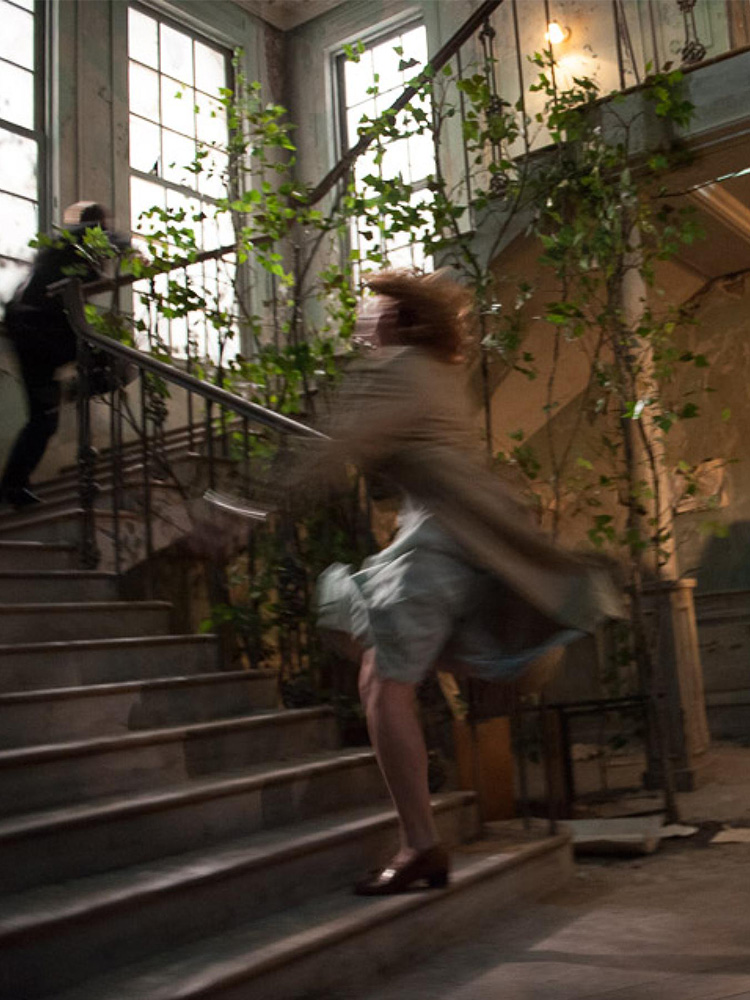 A production photograph of blurred figures running up stairs that have trees or leaves intwined around the bannisters