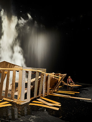 photograph of Phia Ménard’s play Mother House depicting a broken cardboard structure in the foreground, with smoke or dust falling from above the structure, and a figure seated on the floor in the background